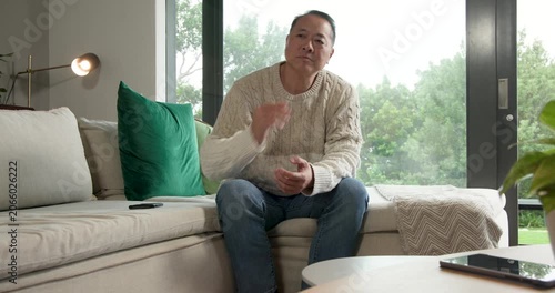 Senior Asian man on sofa, glancing at remote, checking phone, putting it on table, leaving right