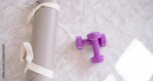 Purple dumbbells are resting on carpet near window as camera is pulling back, revealing rolled mat