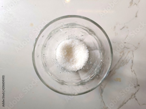 Foto ​A high angle, top down shot of white granulated sugar inside a clear glass cup, placed on a bright marble surface
