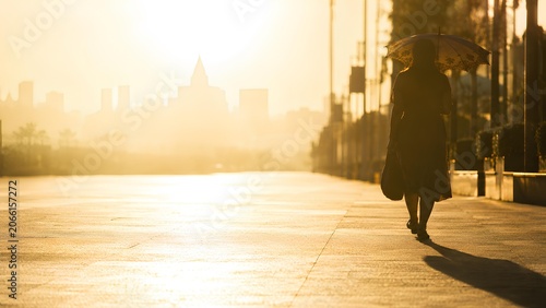 朝日に照らされた都会の遊歩道を歩く傘を差した女性 / Woman walking with umbrella on sunlit city walkway at morning (Generative AI)