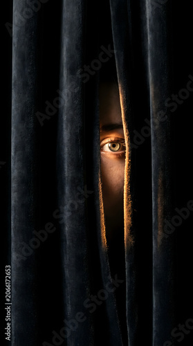 A single human eye peeking through dark velvet curtains with dramatic low light and shadows, conveying a sense of mystery and suspense.
