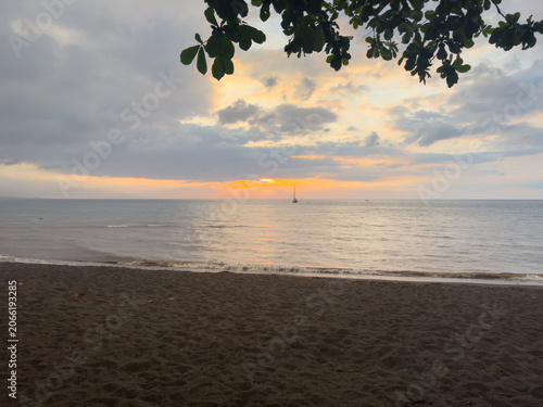 Golden sunset over Lovina Beach in Bali, Indonesia, with silhouetted boat on calm sea and dramatic cloudy sky reflecting on water.