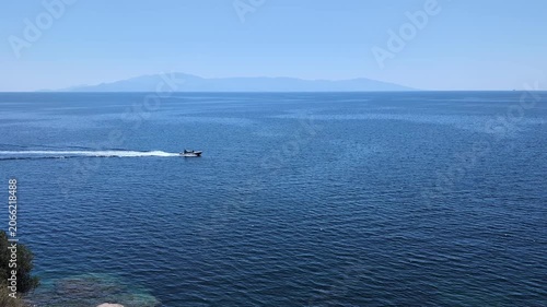 A speedboat speeds across a calm, blue sea under a clear sky, with distant mountains in the background. The water reflects the sunlight, creating a shimmering effect.