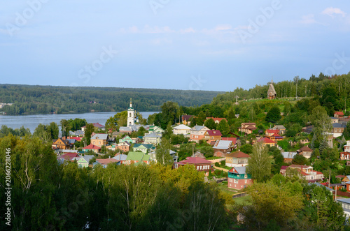 The town of Ples on the banks of the Volga River with wooden houses in a lowland covered with forest, with a church and a bell tower at sunset