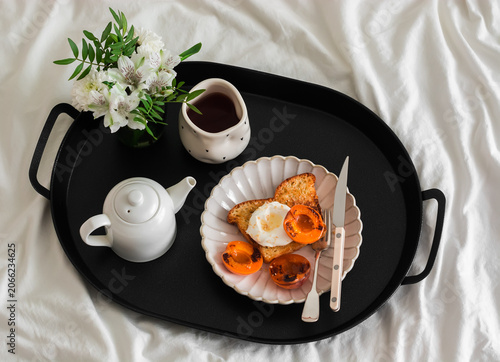 Breakfast in bed - French toast with caramelized apricots, tea and a bouquet of flowers on a black metal tray