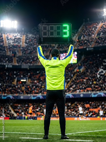 A fourth official in a lime green and blue jacket holds up a digital electronic substitution board showing number three in green LEDs during a night soccer match in a packed stadium.