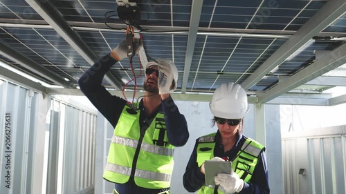 Two solar technicians installing and inspecting microinverter wiring beneath rooftop photovoltaic panels while reviewing system data on a tablet.