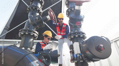Two industrial technicians wearing helmets and safety gear inspecting and adjusting a large valve on mechanical pipeline infrastructure.