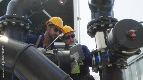 Two industrial technicians wearing helmets and safety gear inspecting and adjusting a large valve on mechanical pipeline infrastructure.