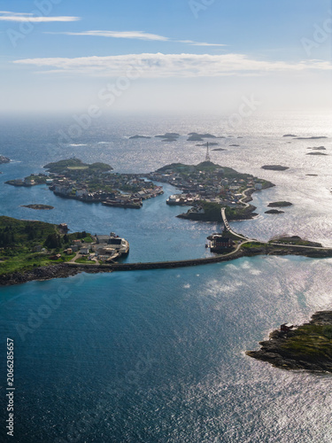 Aerial view of Lofoten near Svolvaer and Henningsvaer, Norway shows rocky islets, turquoise channels, a curving causeway and bridge, small harbors, and clustered buildings on a bright day.