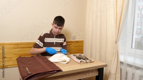 Teenager wearing gloves polishes cutlery with a towel at a table, carefully preparing clean utensils for use.