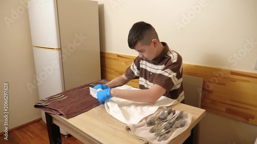 Teenager wearing gloves polishes cutlery with a towel at a table, carefully preparing clean utensils for use.
