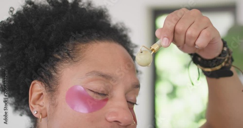 Wearing pink gel patches African American woman rolling tool across brow to soothe skin near window