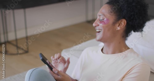 African American woman holding smartphone, tapping, scrolling on light couch at home, relaxing