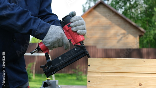 Bild auf Leinwand Close-up of worker in blue uniform and safety gloves using a red pneumatic nail gun to assemble a wooden structure outdoor