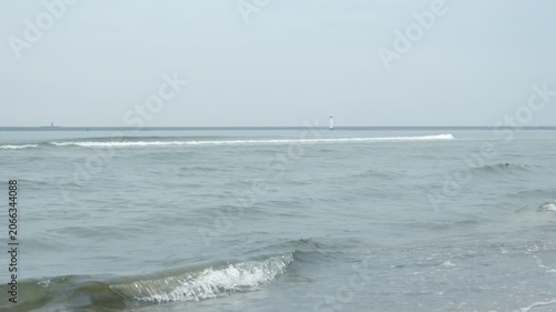 Calm Sea Waves with Overcast Sky and Horizon, Cinematic 4K Video With Audio. Gentle ocean waves rolling onto a sandy beach under a gloomy, overcast sky. The distant misty horizon with lighthouse.