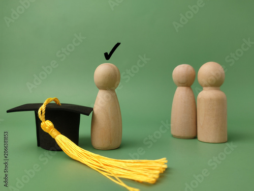 A wooden peg figure stands next to a black graduation cap with a yellow tassel, marked with a check
