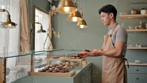 Young confectionery worker placing decorative chocolate figures on shop shelves with pastel interior and hanging lights, handmade sweets display and candy boutique concept