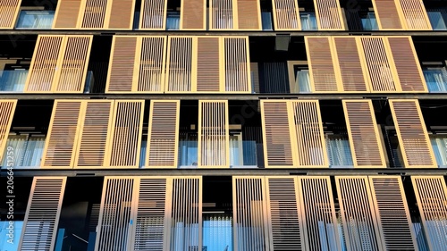 Photo of a contemporary bioclimatic building facade showcasing a rhythmic geometric composition of wooden louvers and sliding sun-shading panels.