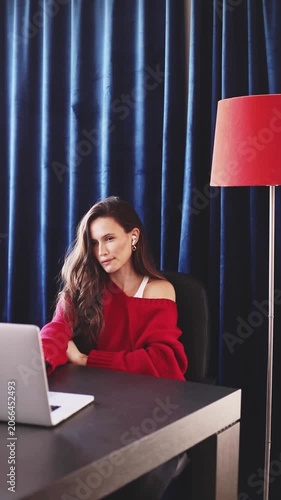 Bored and Tired Woman with Earbuds, Wearing a Red Sweater, Leaning on Her Hand and Sighing While Looking at a Laptop Screen at a Wooden Desk with a Red Floor Lamp and Deep Blue Curtains Background