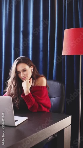 Dreamy and Satisfied Woman in Red Sweater and Earbuds, Smiling Tenderly While Looking at Laptop at Home Office Desk with Red Floor Lamp Against Dark Blue Curtains, Romantic Chat or Movie Content