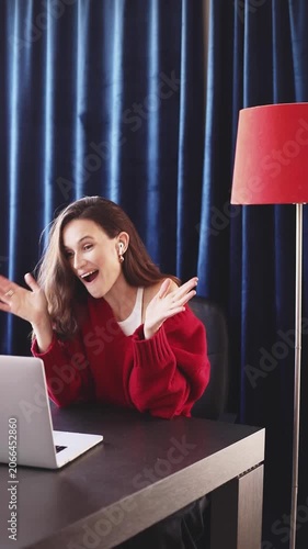 Joyful Woman with Earbuds and Red Sweater, Actively Gesturing and Laughing During Pleasing Video Call on Laptop at Home Office Desk with Red Lamp Against Dark Blue Curtains, Good News Expression