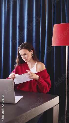 Young Woman at Desk Opening White Envelope, Reading Letter or Important Document with Serious and Focused Expression, Working on Laptop in Home Office with Red Lamp and Blue Curtains Background