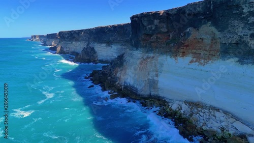Aerial drone shot approaching the sheer Bunda Cliffs from above the ocean with blue sky, blue water, and white waves crashing below