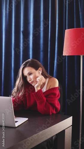 Pensive and Attentive Woman in Red Sweater and Earbuds, Leaning on Hand and Thinking While Working on Laptop at Dark Wooden Desk with Red Floor Lamp Against Blue Curtains, Deep Thinking Concept