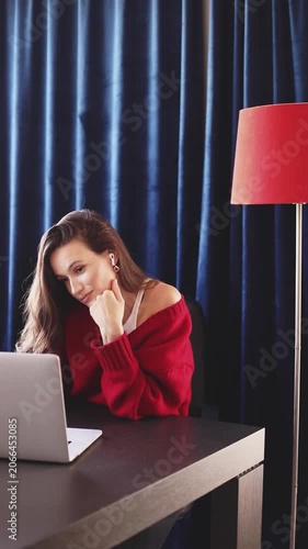 Young Brunette Woman with Earbuds, Wearing Red Sweater, Nodding Her Head as if Agreeing or Listening Attentively to Laptop Screen at Desk with a Red Floor Lamp Against Blue Curtains, Home Office