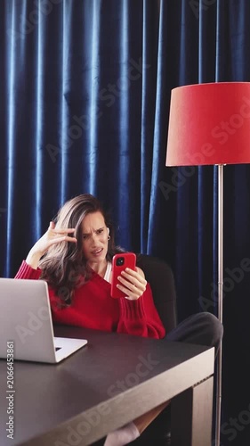 Frustrated and annoyed young woman in red sweater gesturing emotionally while looking at her smartphone, professional remote work challenges, cozy home office, authentic video.