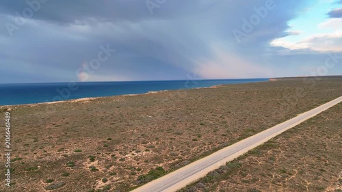 Aerial view following a diagonal road over open wilderness with the Bunda Cliffs, ocean waves, and a bright rainbow on the horizon