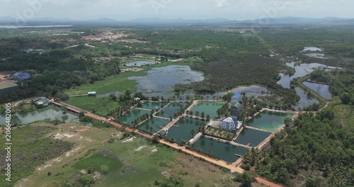 aerial view shows an aquaculture complex with rectangular fish ponds situated in a marshy rural landscape, surrounded by trees and connected by a dirt road.