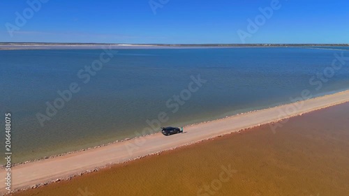 Aerial drone shot orbiting a parked car on the road dividing the pink and blue waters of Point Sinclair Pink Lake, with seagulls flying low over the shimmering surface