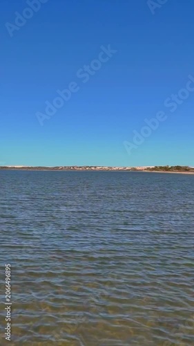 Vertical aerial drone shot flying low over the blue water of Point Sinclair Pink Lake, rising and turning to reveal the pink lake beyond the road