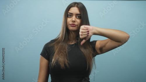 Fotografía Woman with long hair shows thumbs down gesture with hand and visible thumb, pout and frown in a studio blue backdrop; disapproval