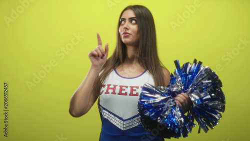 Fényképezés Woman points finger up, mouth open, holding metallic blue pompom, wearing cheer uniform with cheer lettering in studio; team spirit enthusiasm