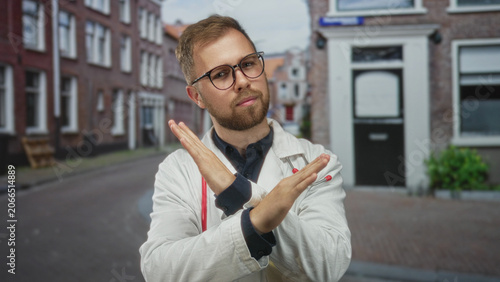Fotografie Man doctor in white coat with stethoscope and glasses crossing arms in street by brick building; professional boundary rejection