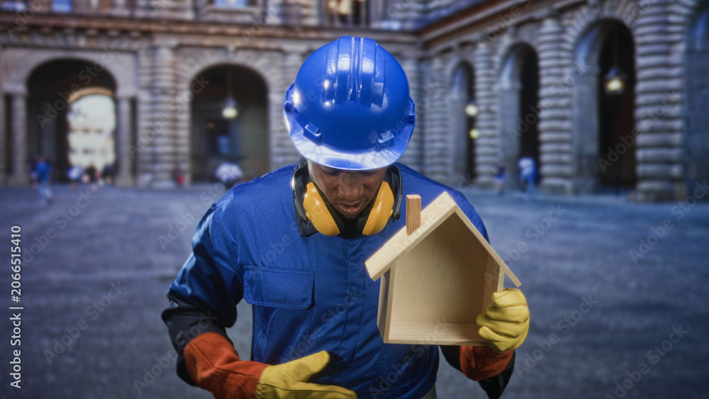 Fototapeta premium Man builder in blue hardhat holds a wooden house model and points with gloved hand in front of building; pride homeownership.
