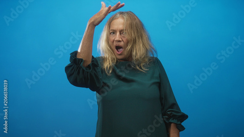 Schilderij op canvas Senior woman with hand on head and open mouth in blue studio, looking startled and gesturing upward; surprise