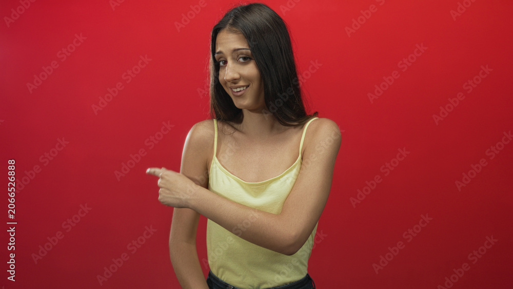 Obraz premium Woman pointing finger to the left in a red studio wearing a yellow tanktop with exposed shoulder and arm; skeptical doubt.