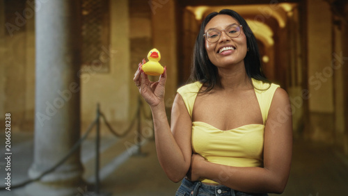 Konstfotografi Woman holding yellow rubber duck while smiling and showing cleavage in ornate building corridor; playful joy