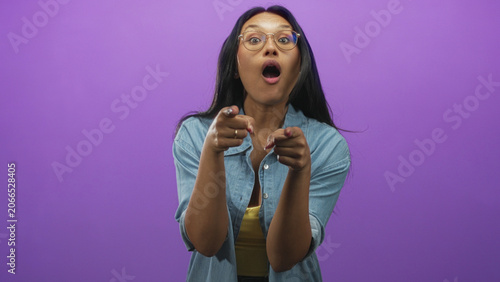 Fotografie Woman pointing both index fingers at camera, smiling, wearing glasses and denim shirt over yellow top in studio; playful confidence