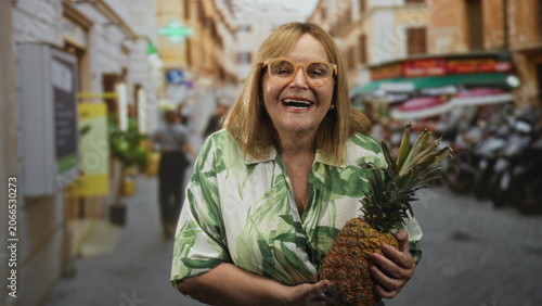 Fotografija Middle age woman holding a ripe pineapple, puckering lips and gesturing with hands on a cobbled street market; playful curiosity