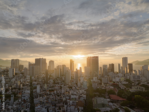 Sunrise above Nha Trang skyline illuminating modern high rise buildings and coastal city streets beneath textured clouds.