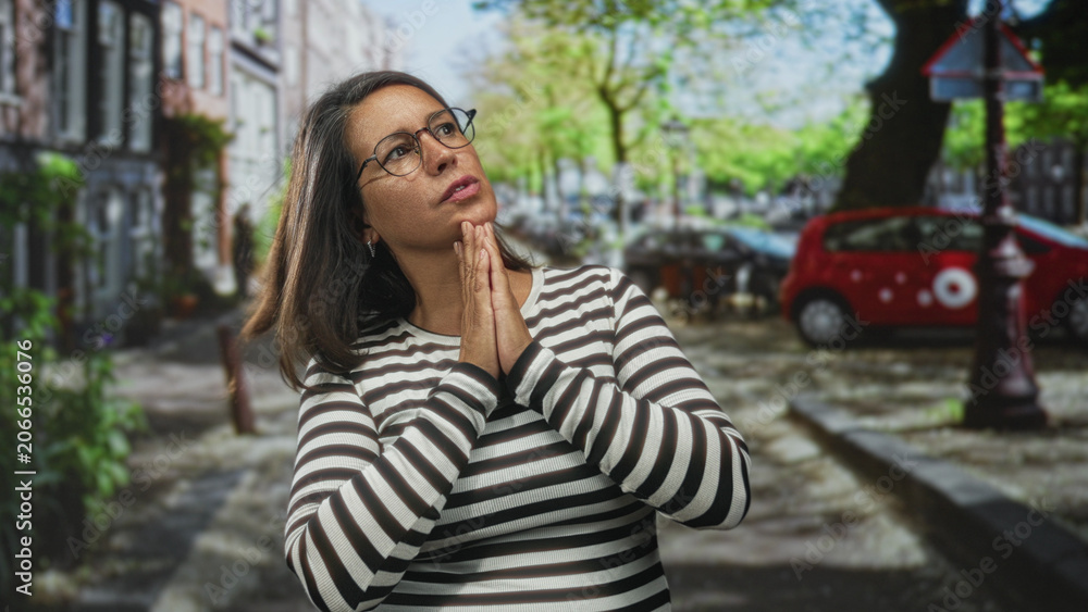 Fototapeta premium Woman brunette middle age clasping hands at chin, wearing glasses and striped top on street with parked car and tree lined lane; quiet contemplation.
