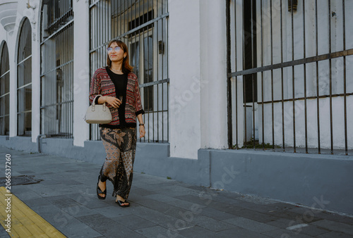 Fashionable adult Asian woman in a glasses, batik textile layered outfit carrying white small bag leather walking on the sidewalk in Surabaya city, Indonesia.