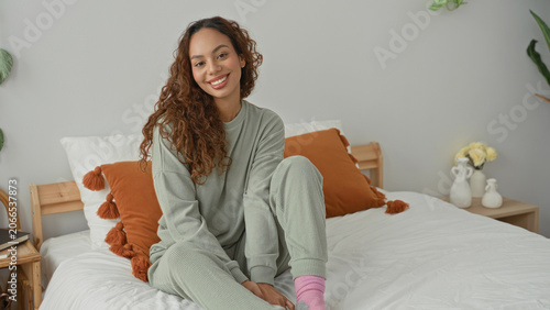 Fotografía Young latin woman in loungewear sitting on bed with hand on knee smiling, curly hair and pink socks, orange pillows visible in building; cozy comfort serene