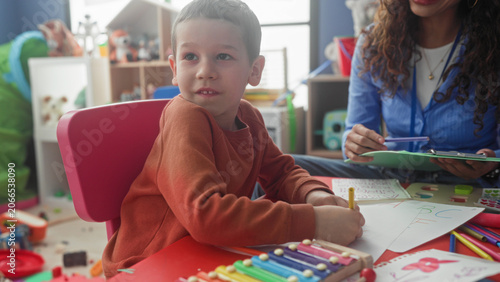 Cuadro en lienzo Boy drawing with pencil using right hand at preschool classroom table while teacher with clipboard watches and smiles; learning focus