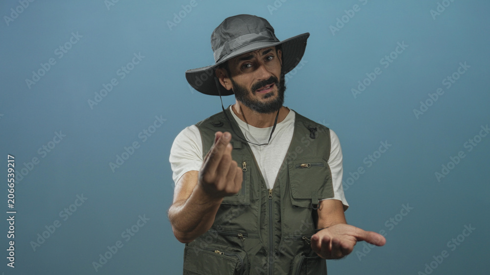 Fototapeta premium Man with beard wearing wide hat and fishing vest beckons with fingers and holds an open hand forward in studio; simple invitation.